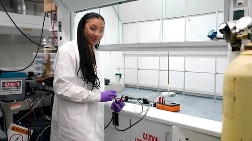 Scientist in safety glasses and purple gloves working with tubing at a lab bench with gas cylinders nearby